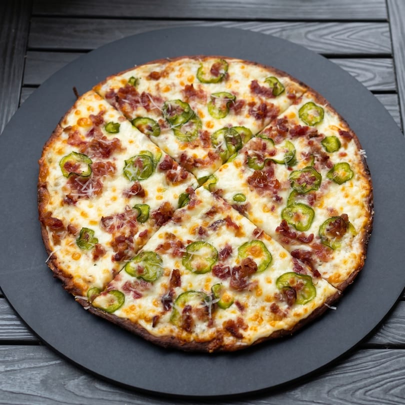 A sliced pizza with melted cheese, bacon bits, and green jalapeño peppers on a dark plate.
