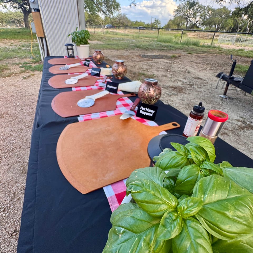 Outdoor pizza-making station with wooden peels, pizza cutters, and fresh basil on a black tablecloth.