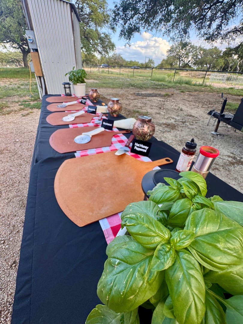 Outdoor pizza-making station with wooden peels, pizza cutters, and fresh basil on a black tablecloth.