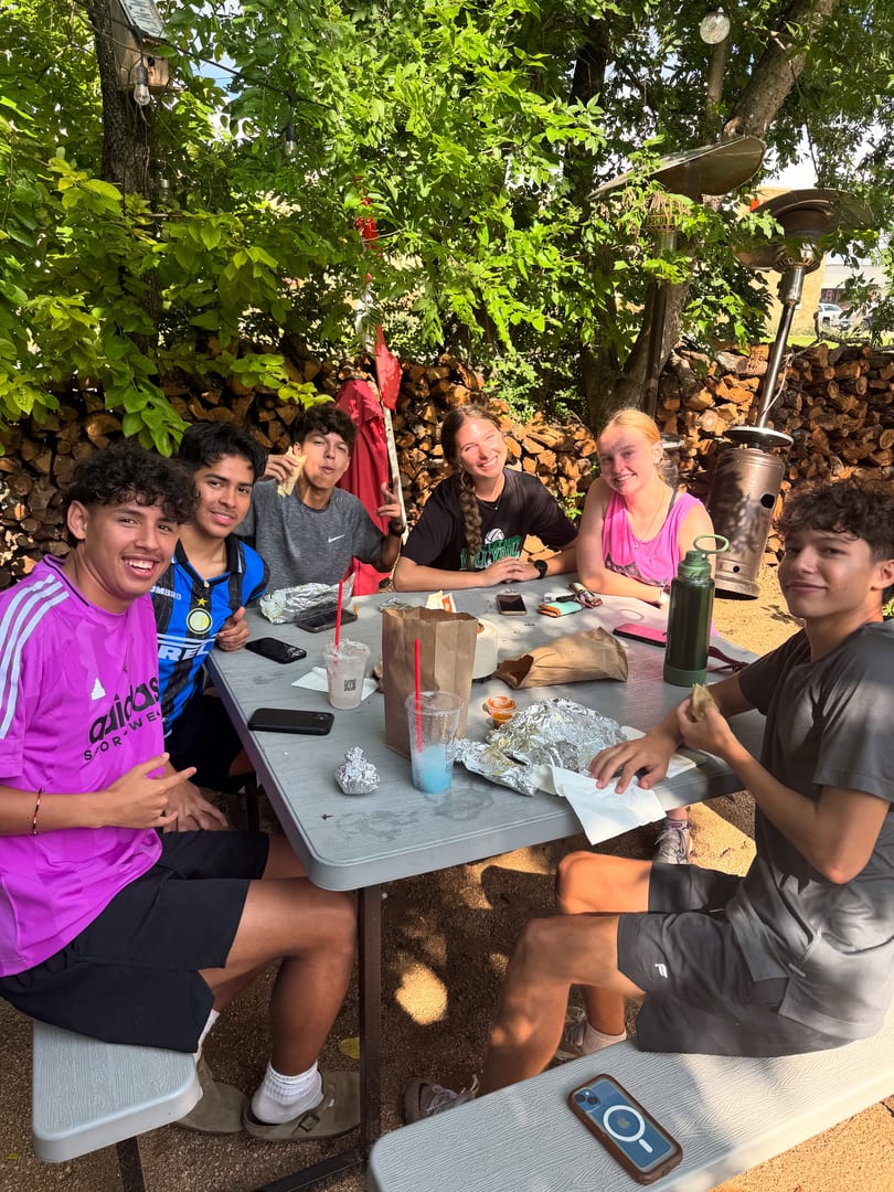 Six young people smiling and eating at an outdoor picnic table under lush green trees.