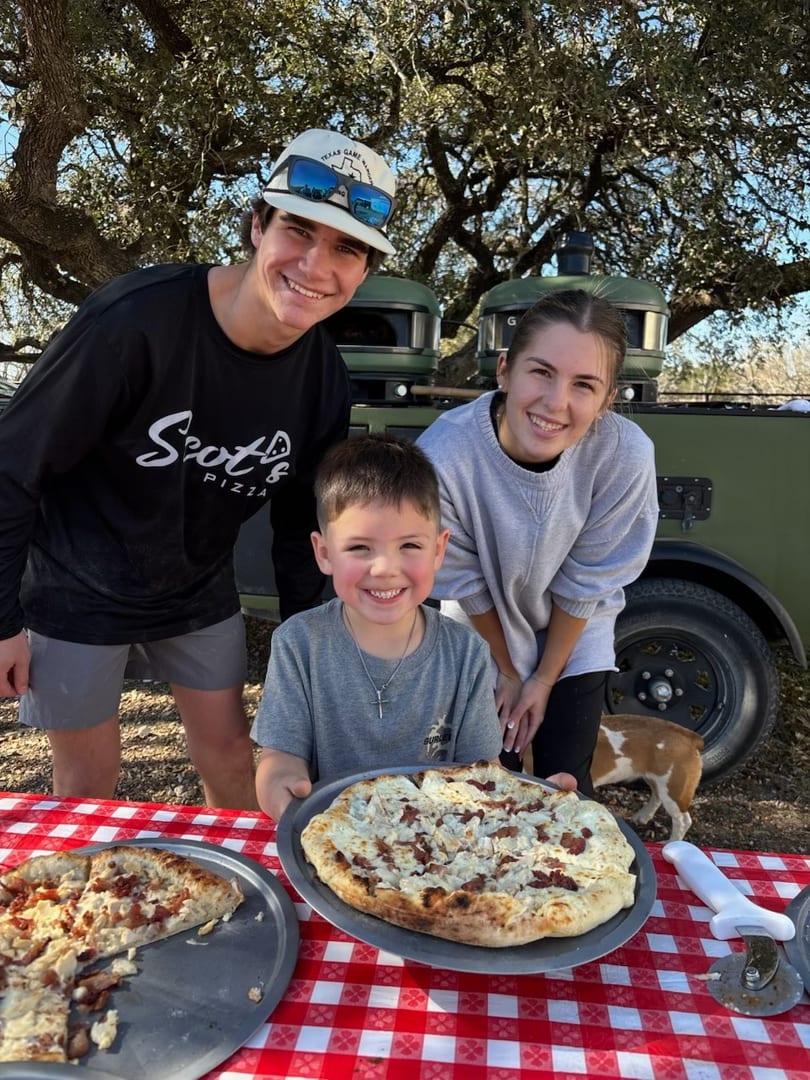 Three smiling people and a dog with two pizzas on a red and white checkered table outdoors.