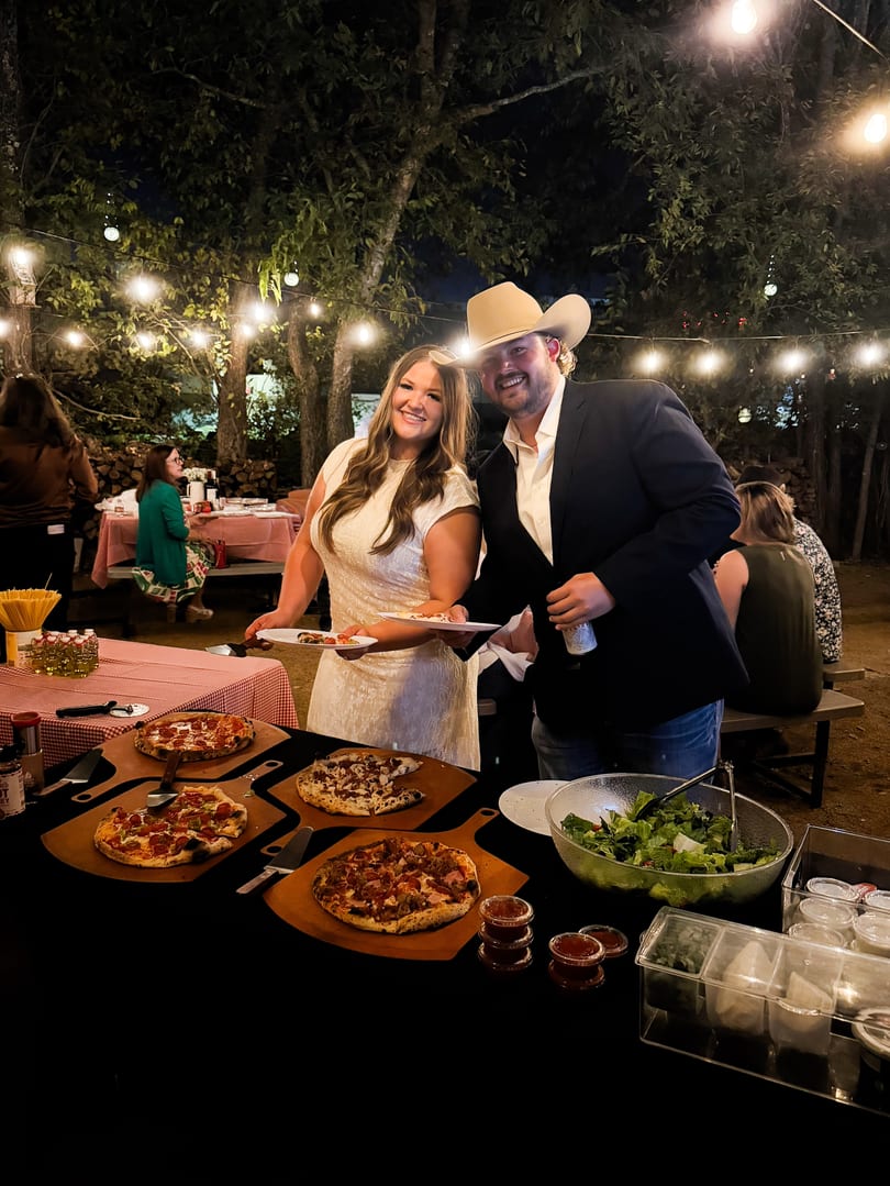A couple smiles while serving themselves pizza and salad from a buffet at an outdoor evening event.