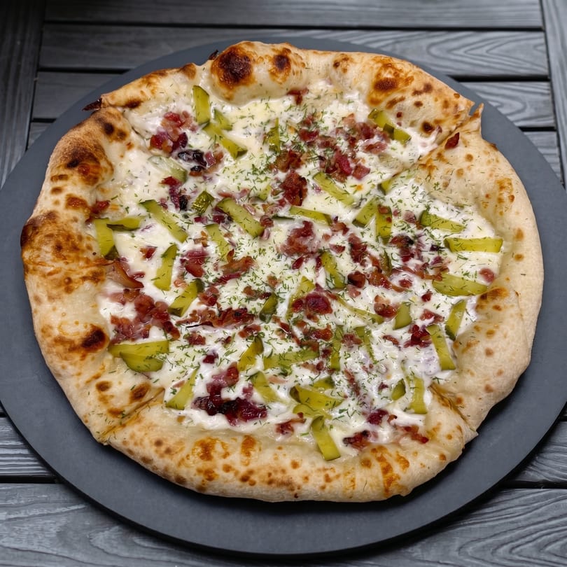 A whole pizza with a white sauce, bacon bits, pickle slices, and dill on a dark plate.
