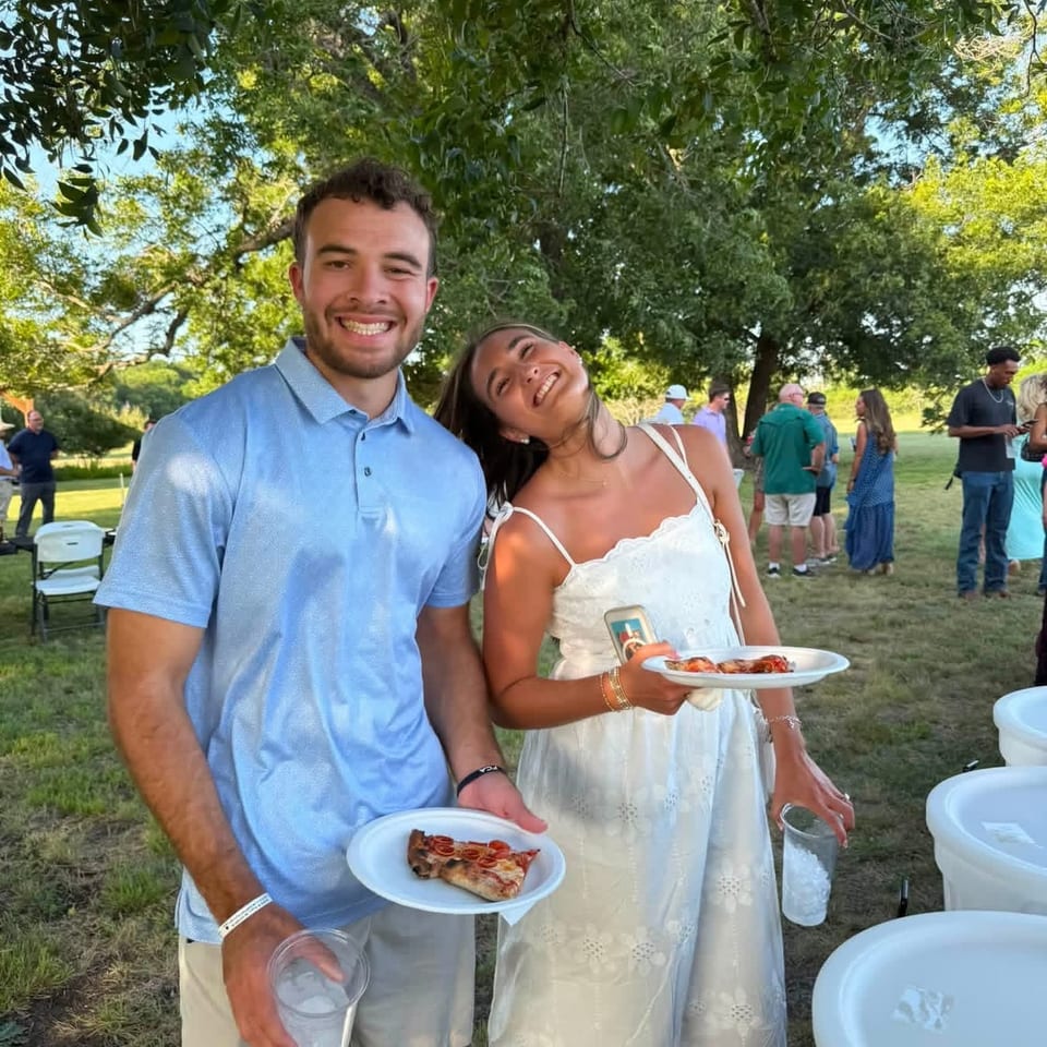 Smiling couple holding plates of pizza and drinks at an outdoor gathering with trees and people in the background.