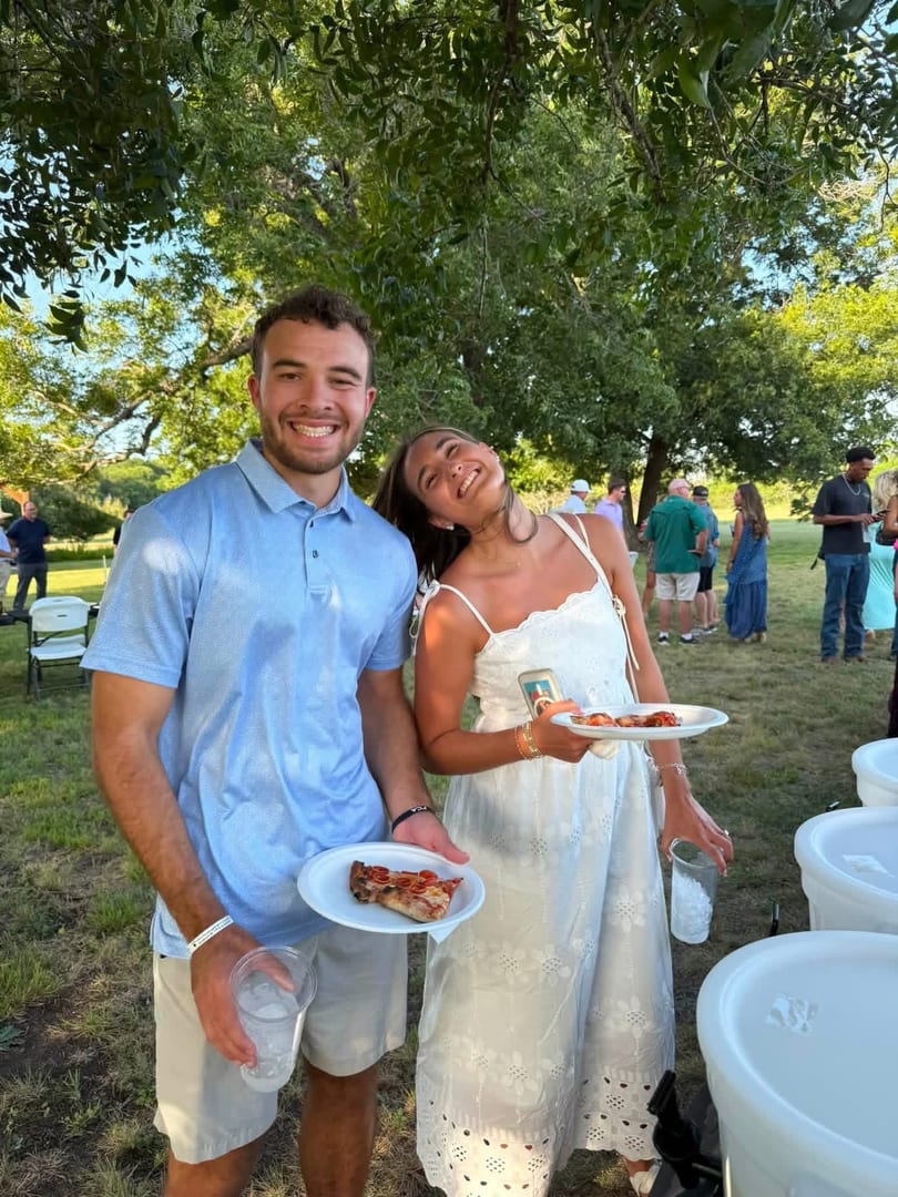 Smiling couple holding plates of pizza and drinks at an outdoor gathering with trees and people in the background.