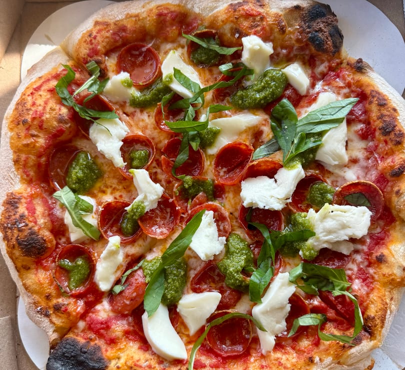 A whole pepperoni pizza with pesto, fresh mozzarella, and basil in a pizza box.