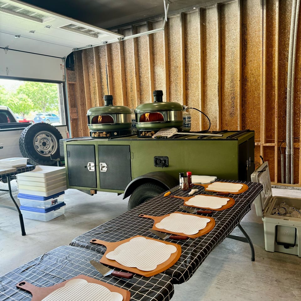 Two green pizza ovens on a trailer, with pizza peels and ingredients on a checkered table.