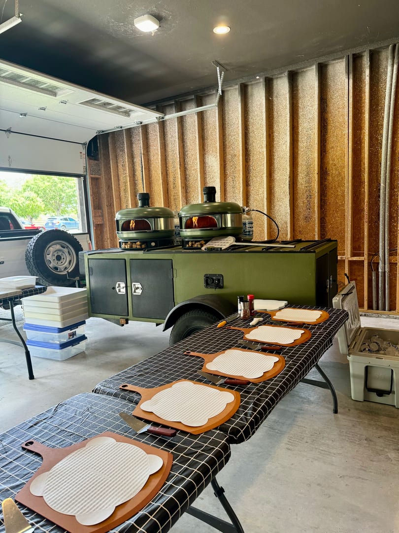 Two green pizza ovens on a trailer, with pizza peels and ingredients on a checkered table.