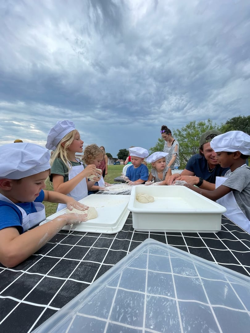 Children in chef hats and aprons happily knead dough at an outdoor table with adults assisting.