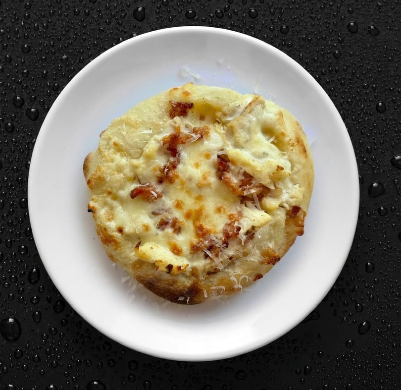 A small, round white pizza with melted cheese and bacon bits on a white plate.