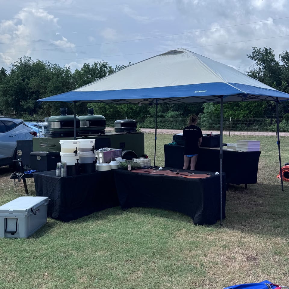 Outdoor catering setup with a blue and white tent, food tables, and a person preparing food.