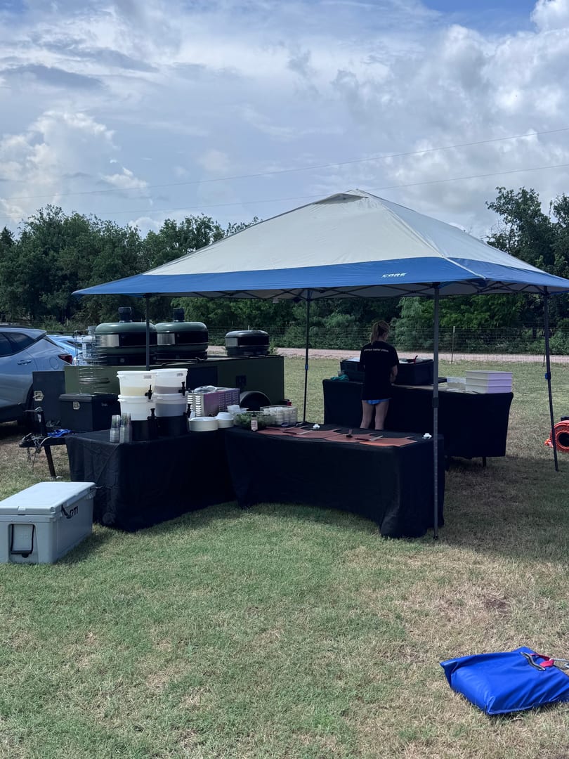 Outdoor catering setup with a blue and white tent, food tables, and a person preparing food.