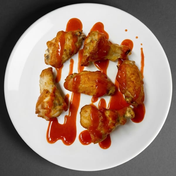Six golden-brown chicken wings drizzled with red hot sauce, arranged on a white plate.
