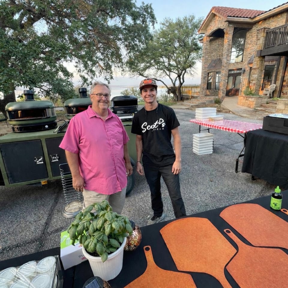 Two men stand beside pizza ovens and a table with basil and pizza peels, outside a stone house.