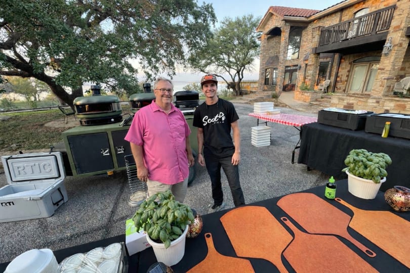 Two men stand beside pizza ovens and a table with basil and pizza peels, outside a stone house.