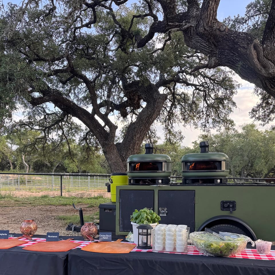 Outdoor pizza ovens and a buffet table with pizza ingredients and salads under large oak trees.