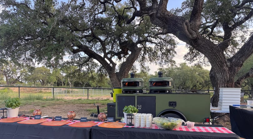 Outdoor pizza ovens and a buffet table with pizza ingredients and salads under large oak trees.