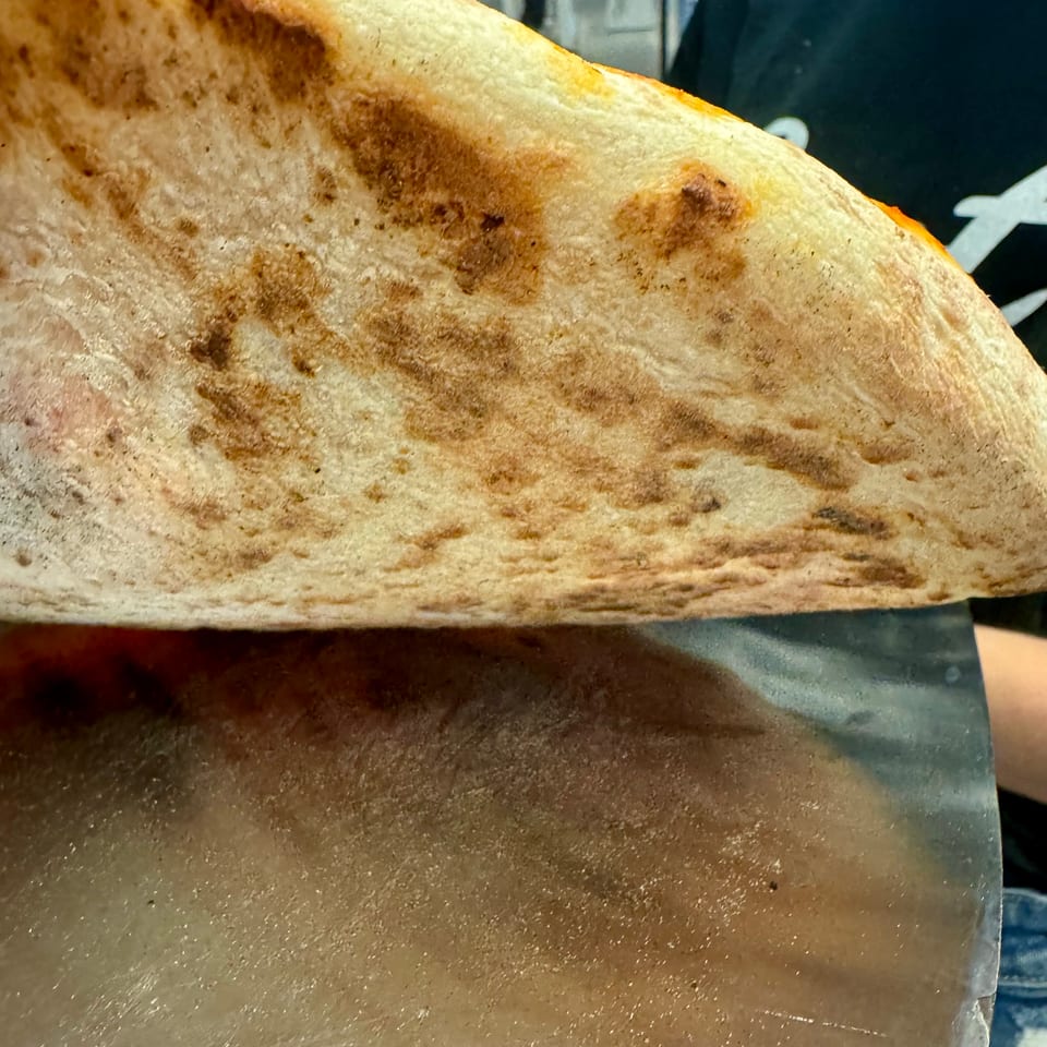 Close-up of a pizza crust with charred spots being lifted by a metal peel.