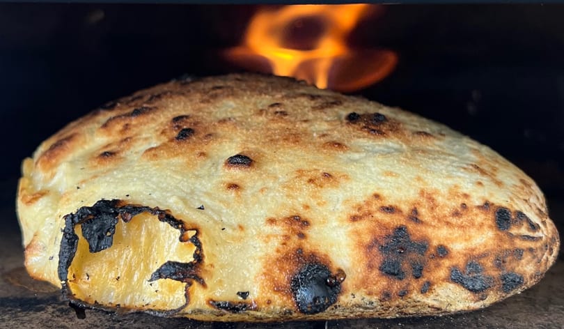 A calzone with a golden-brown crust and charred edges baking in a wood-fired oven.