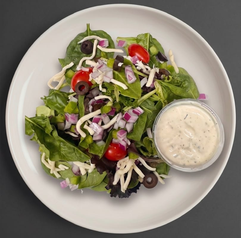 Fresh garden salad with mixed greens, tomatoes, olives, onions, cheese, and a side of dressing.