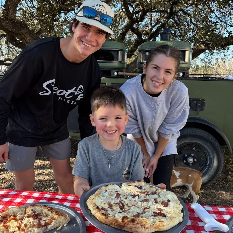 Three smiling people and a dog with fresh pizzas on a red and white checkered table outdoors.