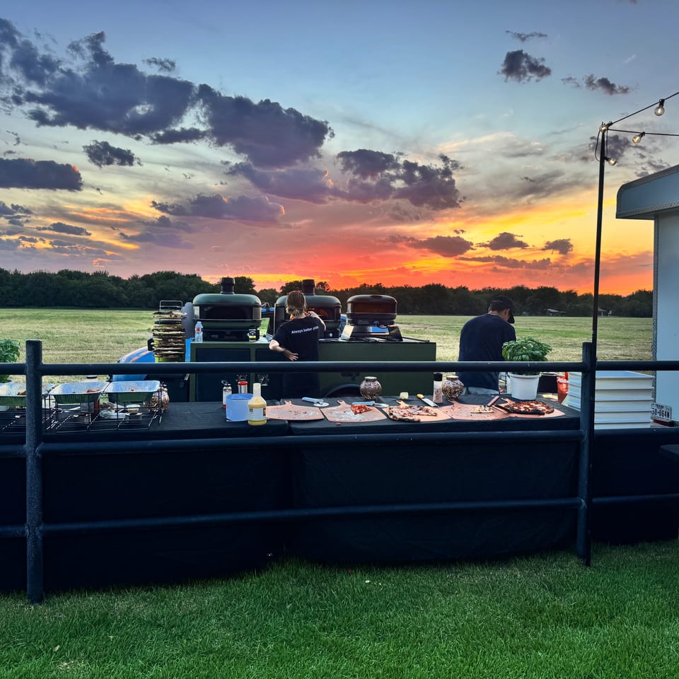 Outdoor catering setup with pizza ovens and food tables under a vibrant sunset sky.