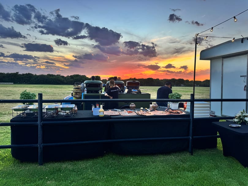 Outdoor catering setup with pizza ovens and food tables under a vibrant sunset sky.
