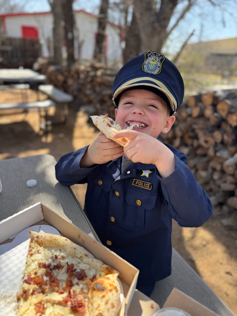 A young boy in a police costume happily eats a slice of pizza outdoors.