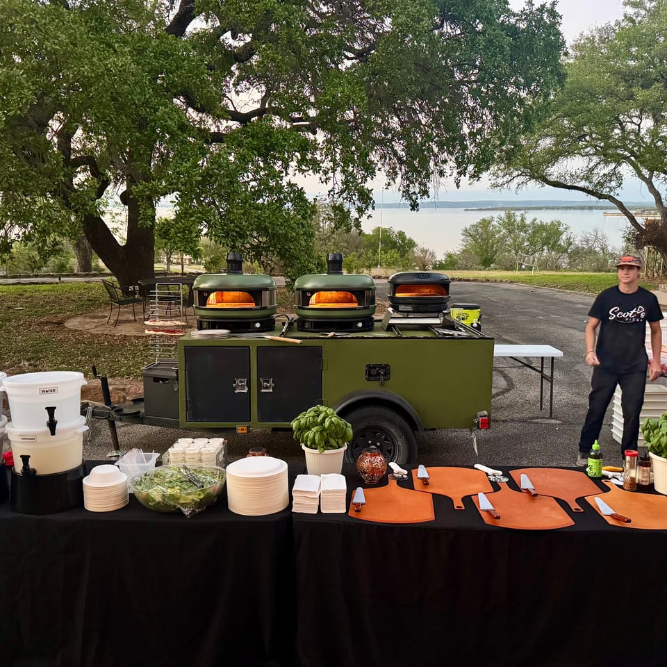 A mobile pizza kitchen with three ovens, a beverage station, and a serving table outdoors by a lake.