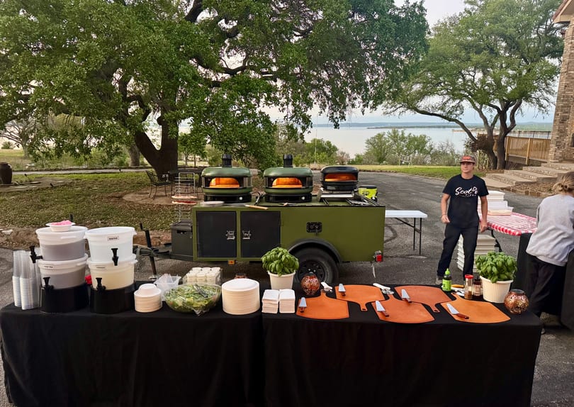 A mobile pizza kitchen with three ovens, a beverage station, and a serving table outdoors by a lake.